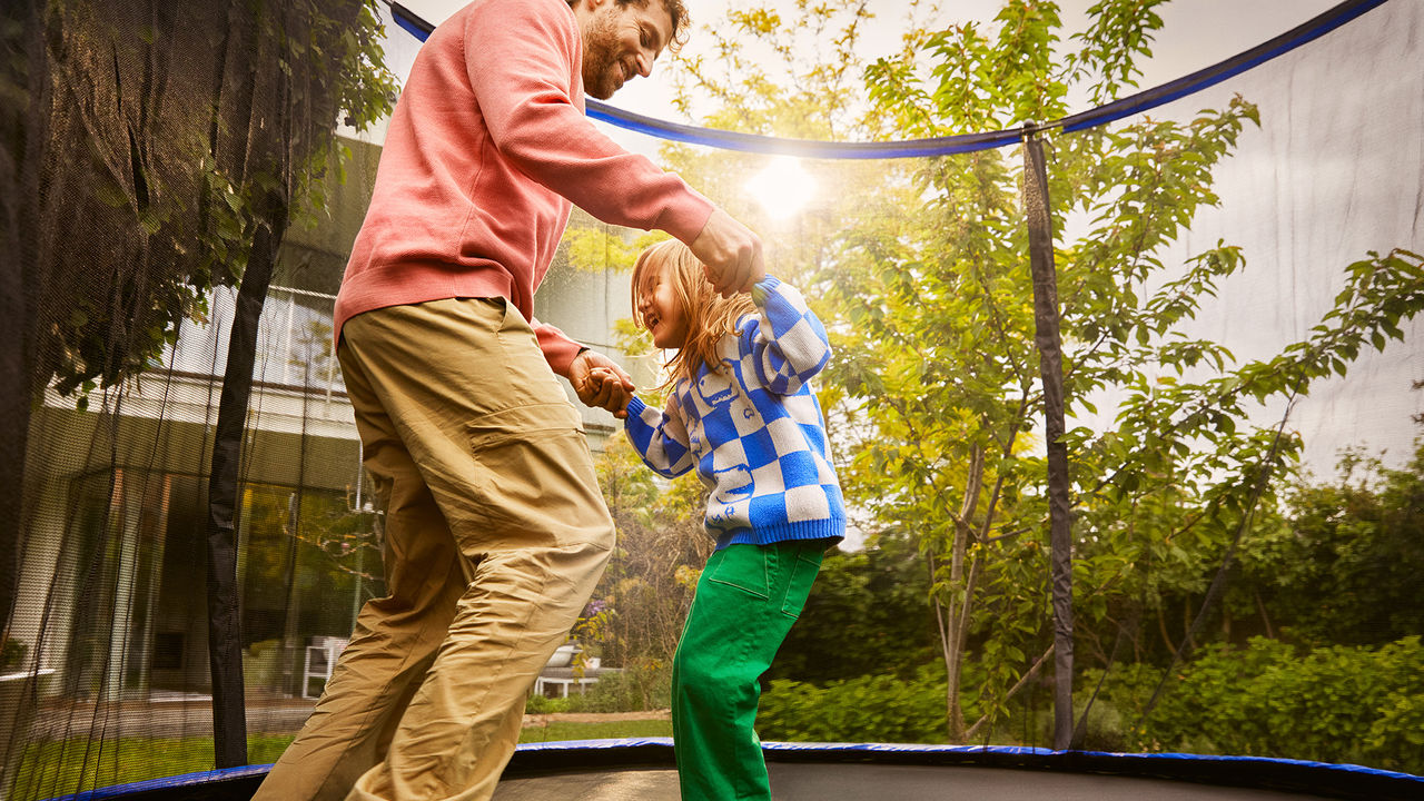 Ein Vater springt gemeinsam mit seiner Tochter im Garten auf einem Trampolin.