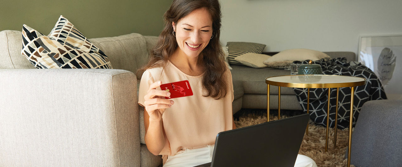 Eine junge Frau sitzt in ihrem Wohnzimmer und verwendet ihre Wüstenrot Karte um einen online Einkauf abzuschließen.