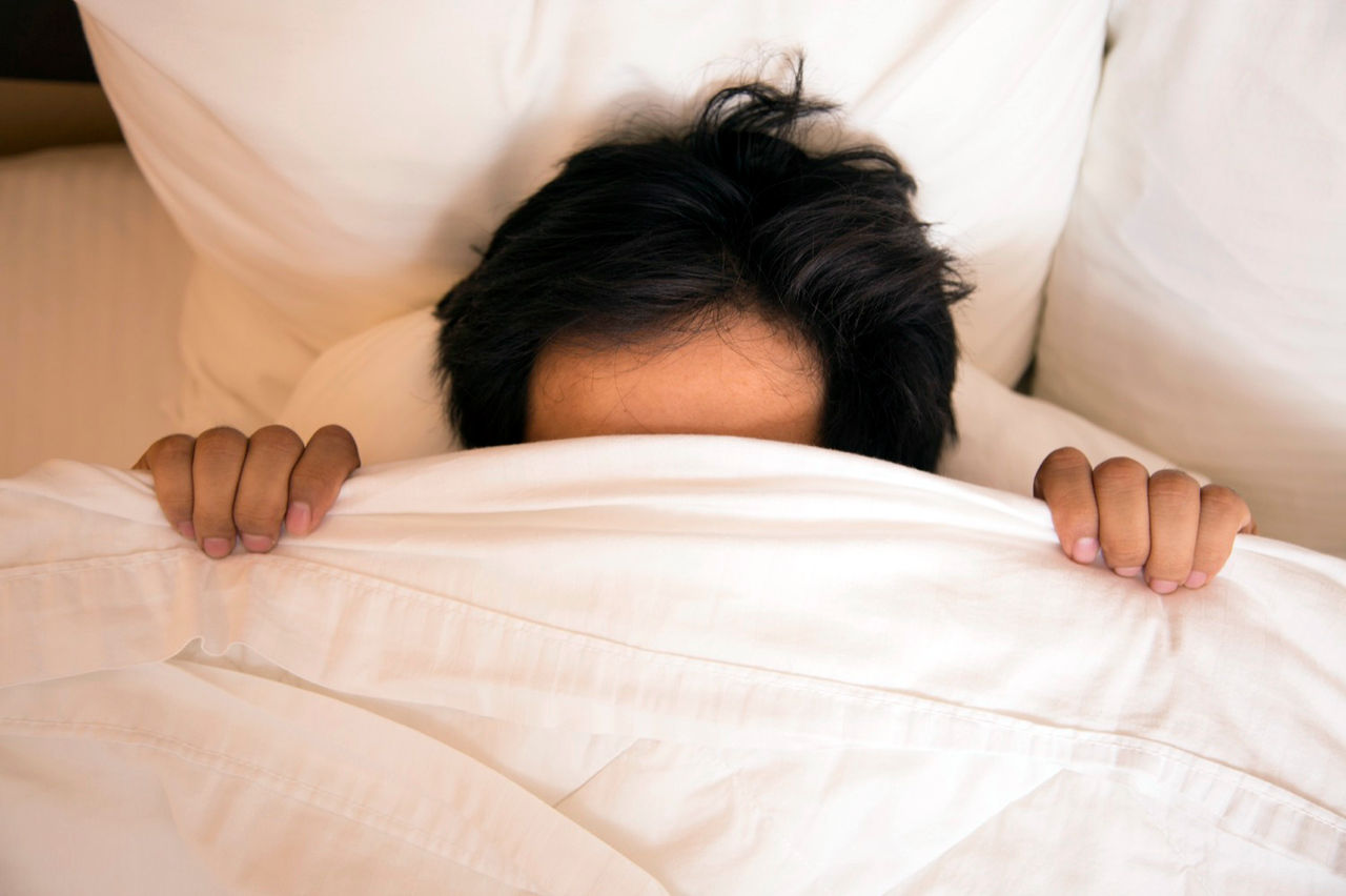 Do not disturb.  Asian descent man pulls up the blanket over his head in his hotel room bed.  He is fast asleep with his head on soft, fluffy pillows in a comfortable hotel bed. Only his forehead and top of head are showing.  Pillows and sheets around him. Luxury room. Great copyspace in blankets.