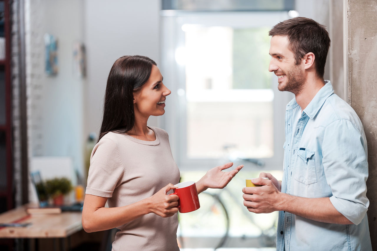 Two cheerful young people holding coffee cups and talking while standing in office