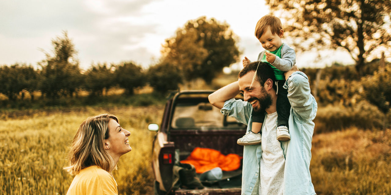 Mobilitätskasko - Auto Kaskoversicherung - Paar mit Kind - Vater mit Kleinkind auf den Schulter und Auto im Hintergrund 